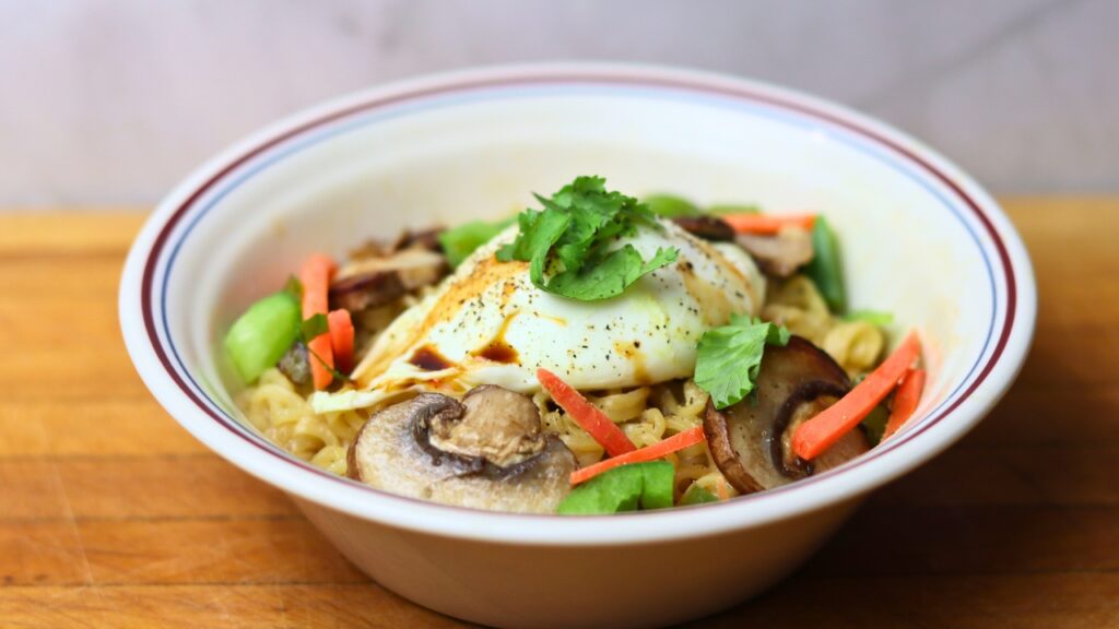 Bowl of ramen noodles topped with veggies and a poached egg garnished with parsley on a wooden countertop.