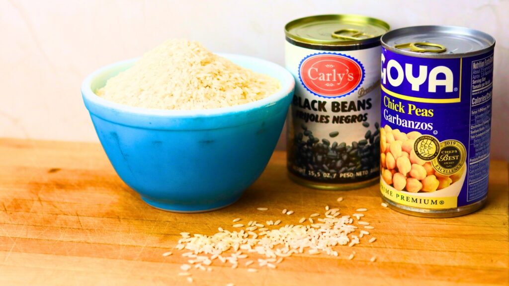 Blue glass bowl of white rice next to two cans of beans on a wooden counter top for cheap pantry meals.