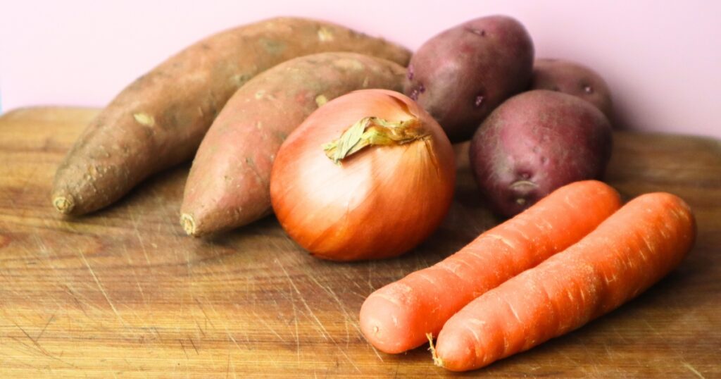 Sweet potatoes, red potatoes, a yellow onion and two orange carrots on a wooden sideboard.